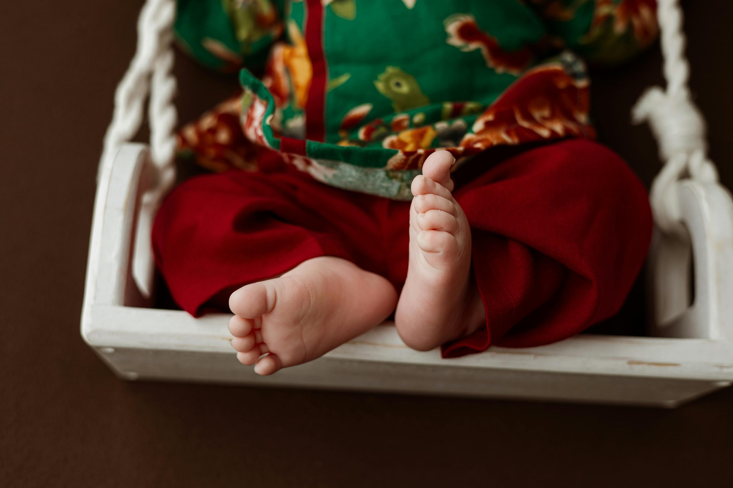 Adorable baby feet in traditional outfit resting on a wooden swing.